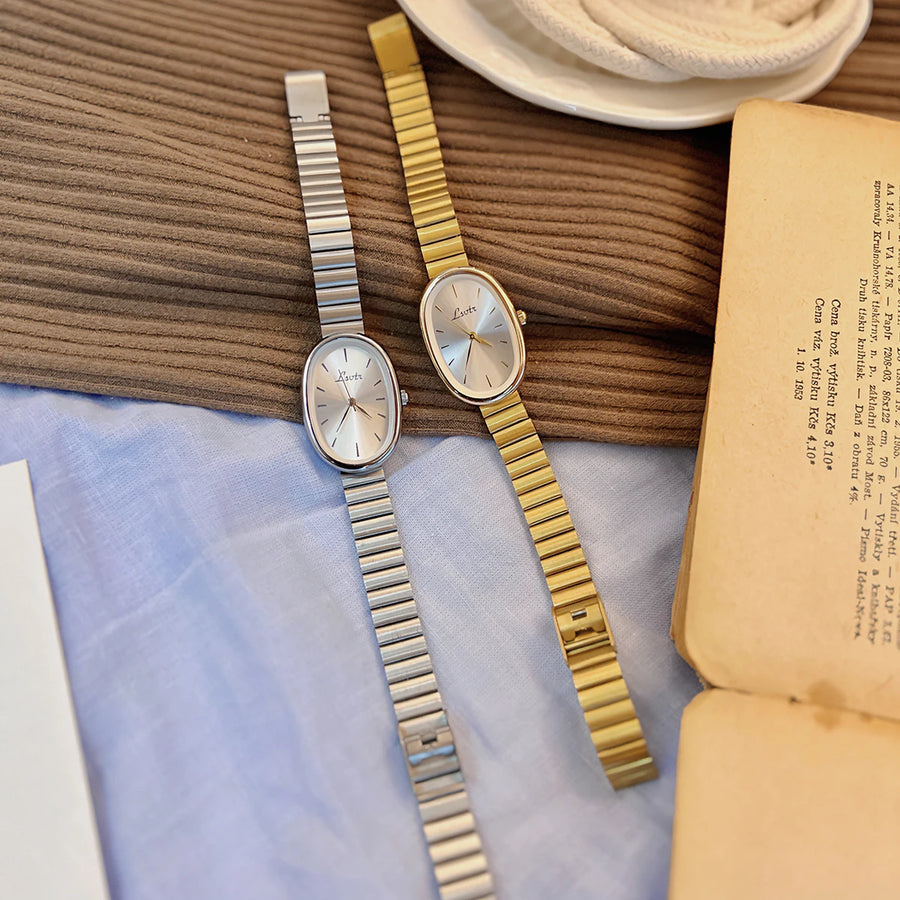 Two watches, one silver and one gold, on a textured surface with an open book.