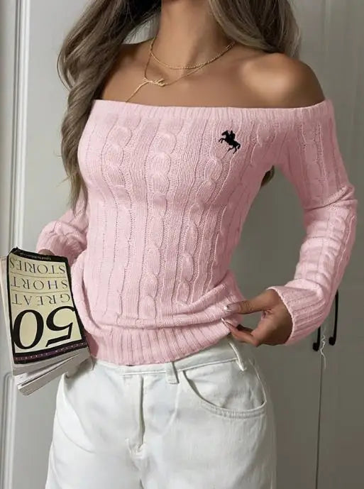 Model wearing pink off-shoulder cable-knit sweater with embroidered logo, holding a book