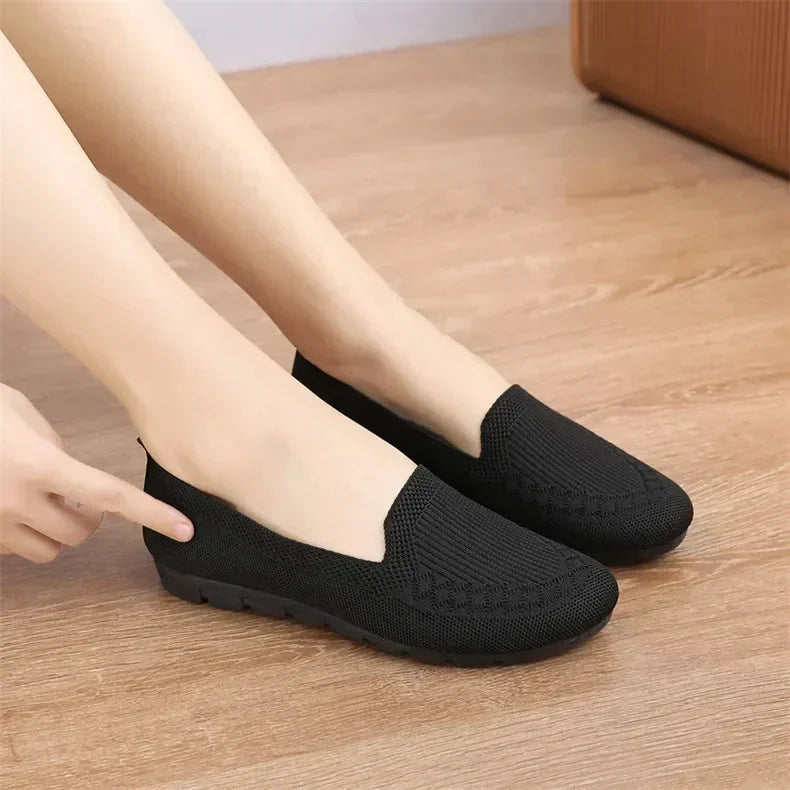 Black slip-on shoes worn by a person on a wooden floor