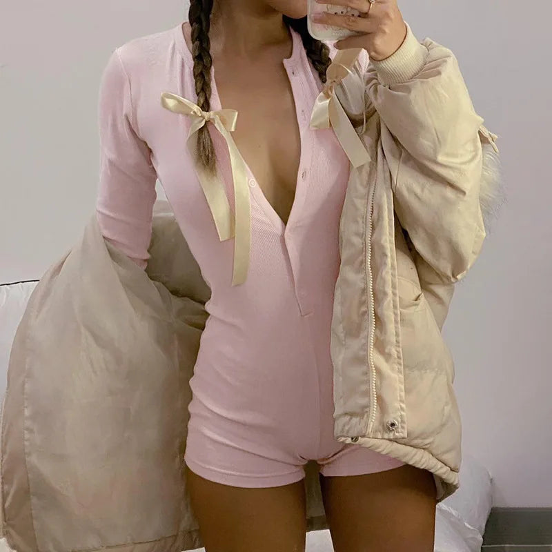 Model taking a selfie wearing pink long sleeve button-front romper with cream ribbons in braided hair, holding a beige puffer jacket