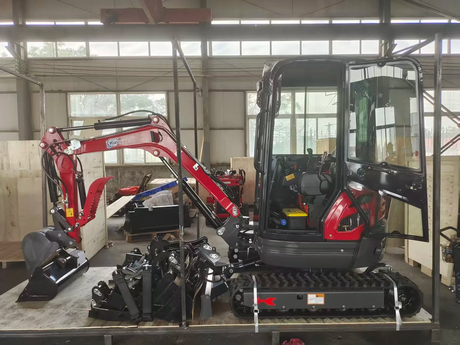 A small red and black crawler excavator with an open cab and attached bucket is photographed inside a warehouse or workshop. Metal support frames and other equipment are visible around it, and large windows and industrial ceiling structure are in the background.