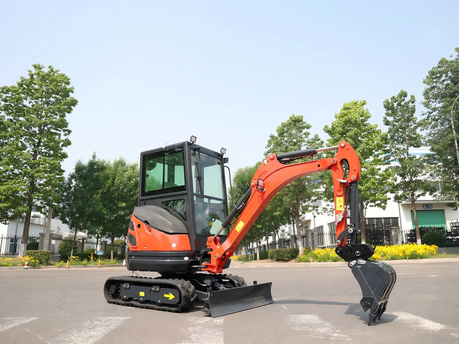 An orange and black compact crawler excavator with a front blade and bucket attachment, photographed outdoors on a paved area with trees and buildings in the background.