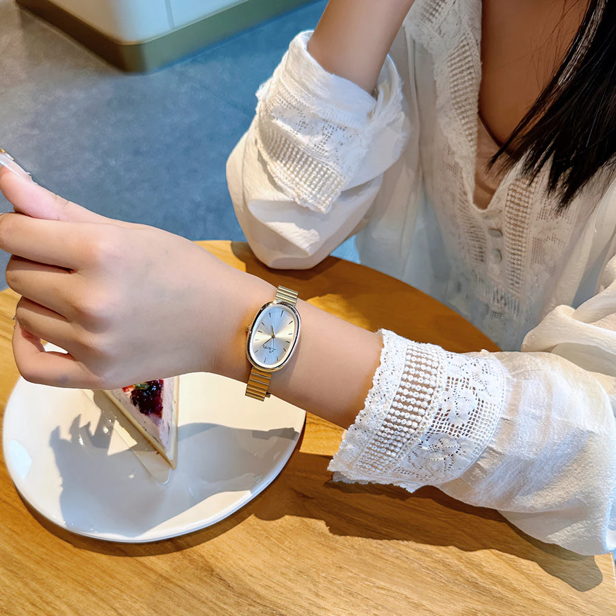 Person wearing a gold watch on a wooden table with a blurred background