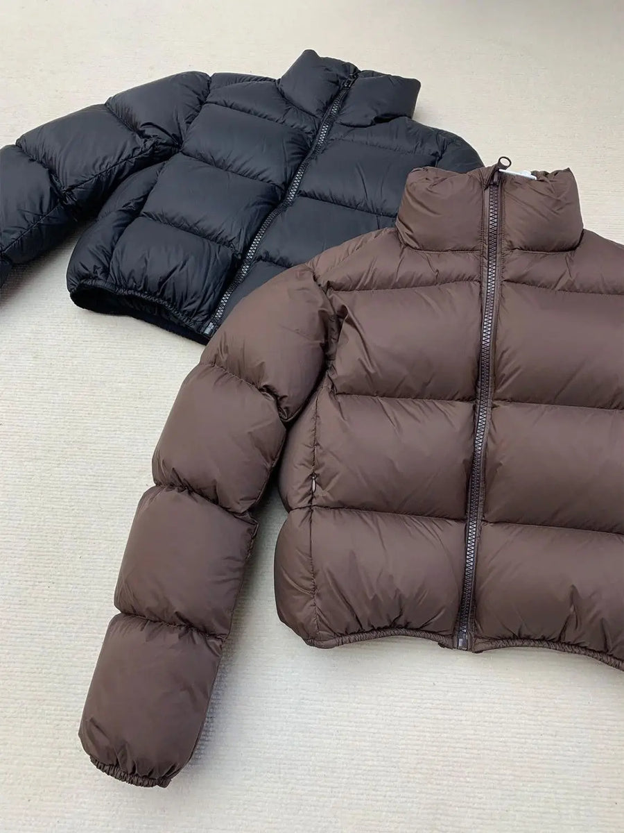 Two cropped puffer jackets, one black and one brown, on a light gray surface