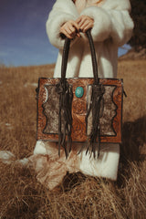 Person holding a decorative handbag with intricate patterns and a turquoise stone in a field.