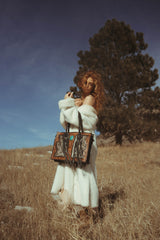 Woman in a white fur coat holding two patterned handbags in a field with a tree in the background.