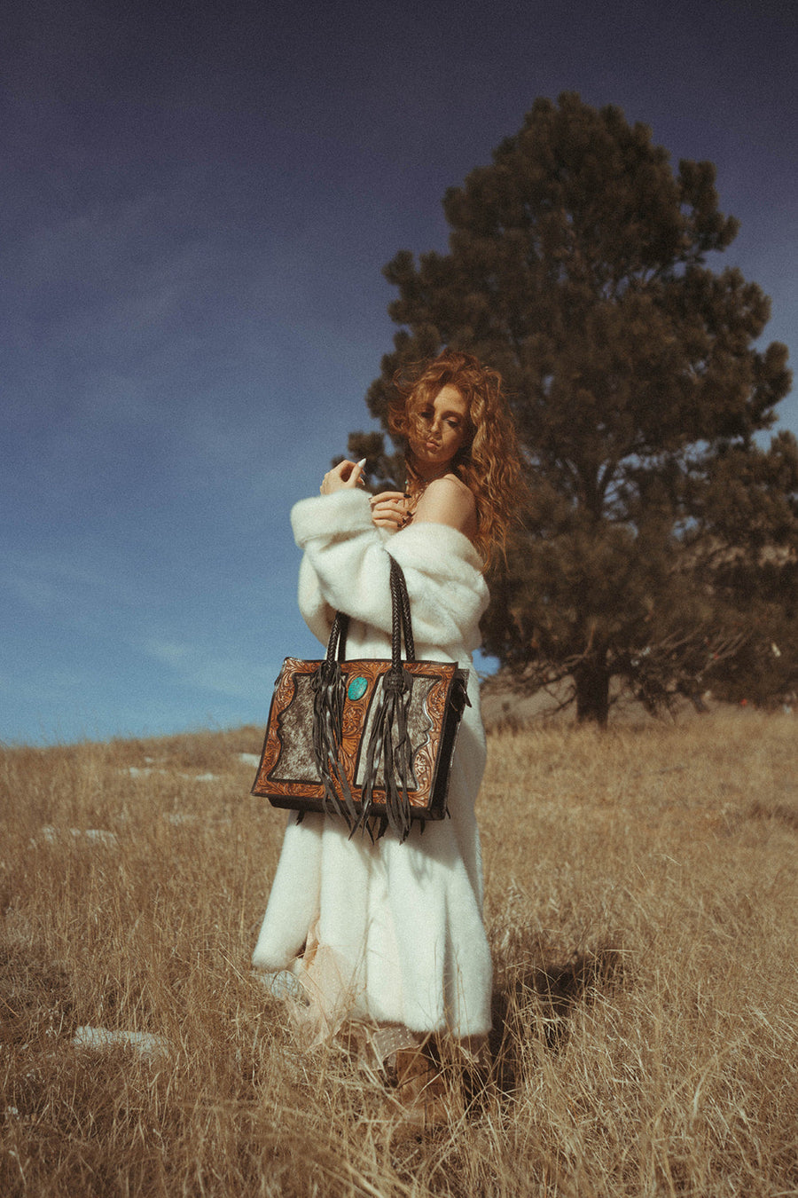 Woman in a white fur coat holding two patterned handbags in a field with a tree in the background.