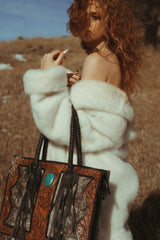 Woman in a white fur coat holding a decorative brown bag with a turquoise stone in a natural setting.