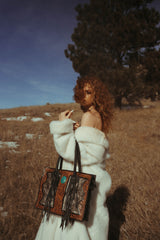 Woman in a white fur coat holding a decorative handbag in a field with trees in the background