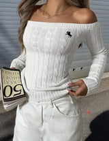 Model wearing white off-shoulder cable-knit sweater with embroidered horse logo and white pants, holding a book