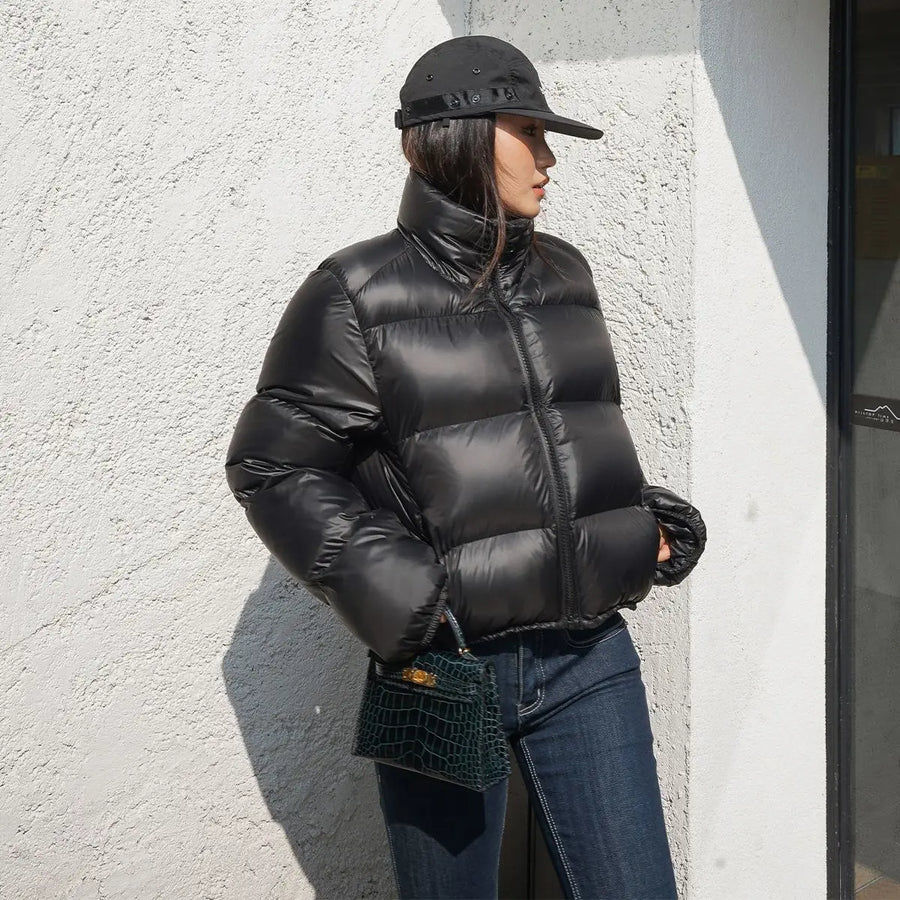 Person wearing a black cropped puffer jacket and cap against a light-colored wall