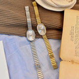 Two watches, one silver and one gold, on a textured surface with an open book.