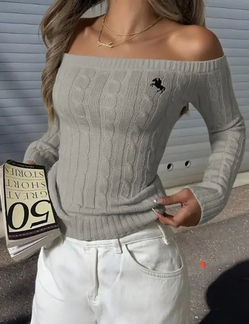 Model wearing light gray off-shoulder cable-knit sweater with embroidered horse logo, holding a book and wearing white pants