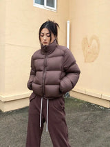 Person wearing a brown cropped puffer jacket and pants standing against a beige wall