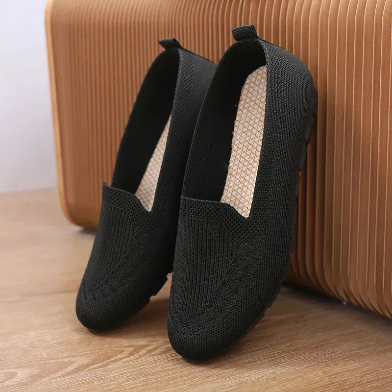 Pair of black knit shoes on a wooden floor with a textured brown wall in the background.