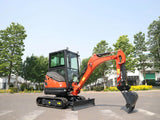 An orange and black compact crawler excavator with a front blade and bucket attachment, photographed outdoors on a paved area with trees and buildings in the background.
