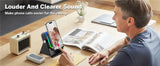 Person using a device with a phone on a stand, sitting at a table with a magazine and small clock.