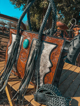 Handcrafted leather bag with turquoise stone and fringe details on a wooden surface.