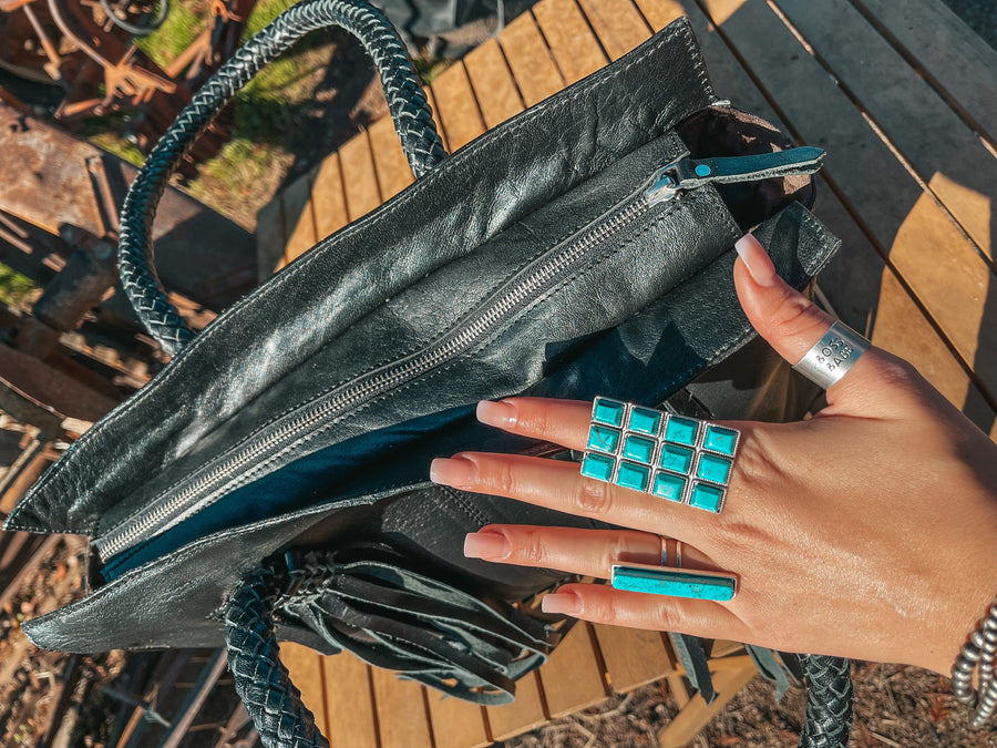 Hand with turquoise ring opening a black handbag on a wooden surface