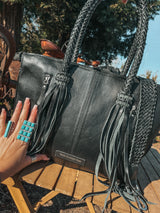 Black handbag with braided handles held by a hand, outdoors.